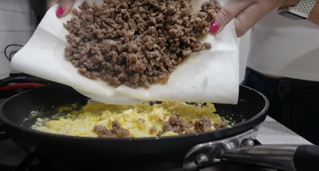 Lindsay is pouring the cooked ground turkey meat into the pan with the scrambled eggs